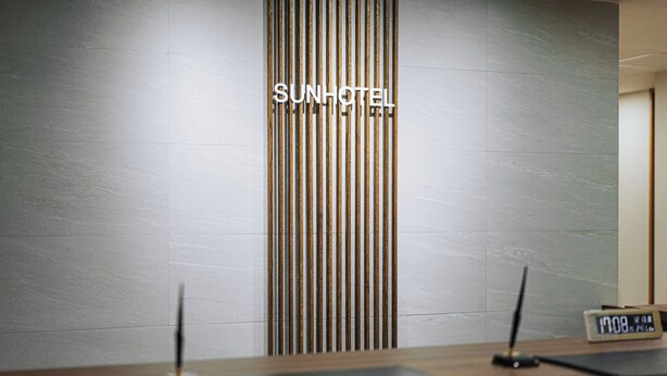 A reception desk with a modern design, featuring a vertical wooden panel and a digital clock displaying the time. - Business Inn SUNHOTEL (Machida)
