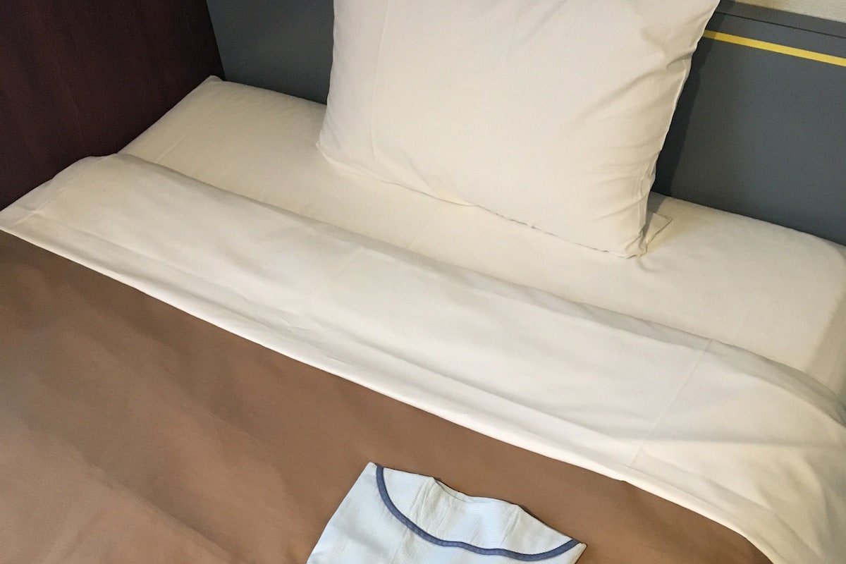 A bed with a white sheet, a brown blanket, and a folded white shirt with a blue collar.