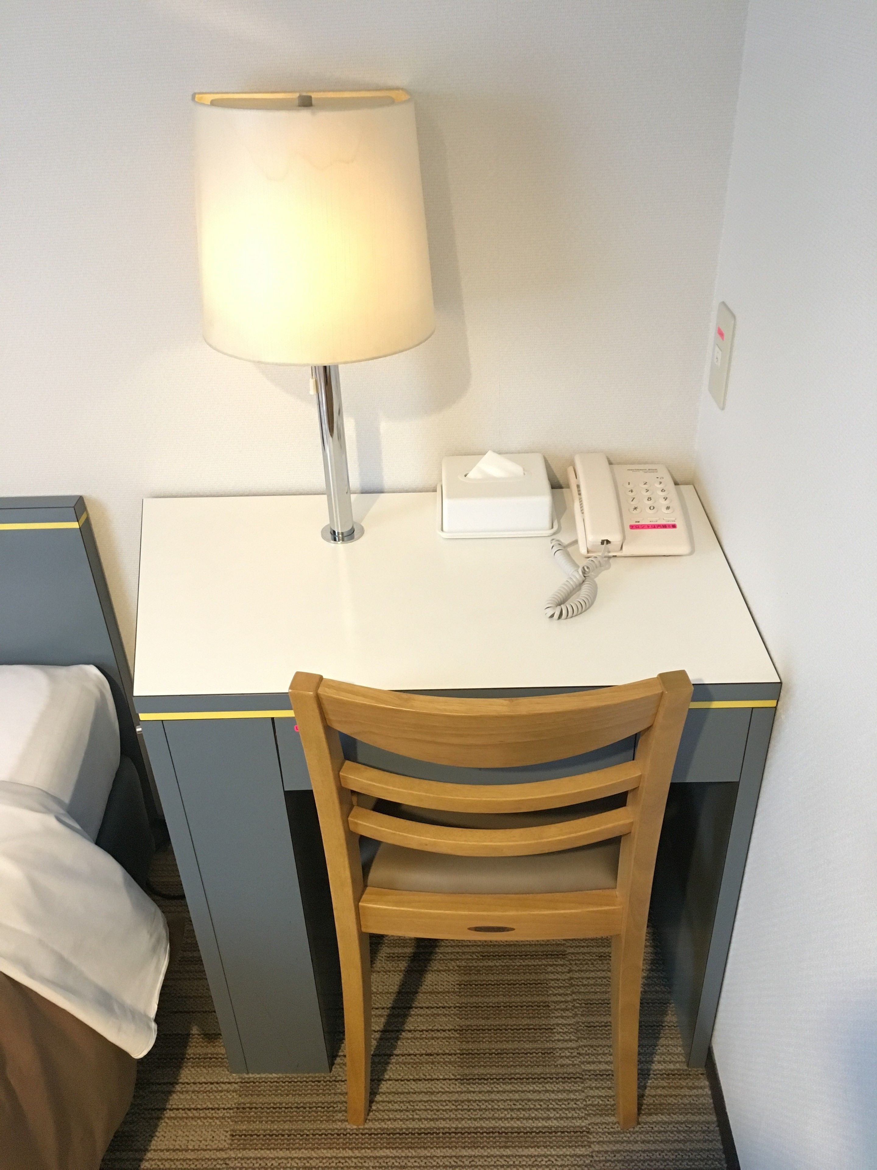 A hotel room with a desk, a telephone, a lamp, a chair, and a bedside table with a tissue box.