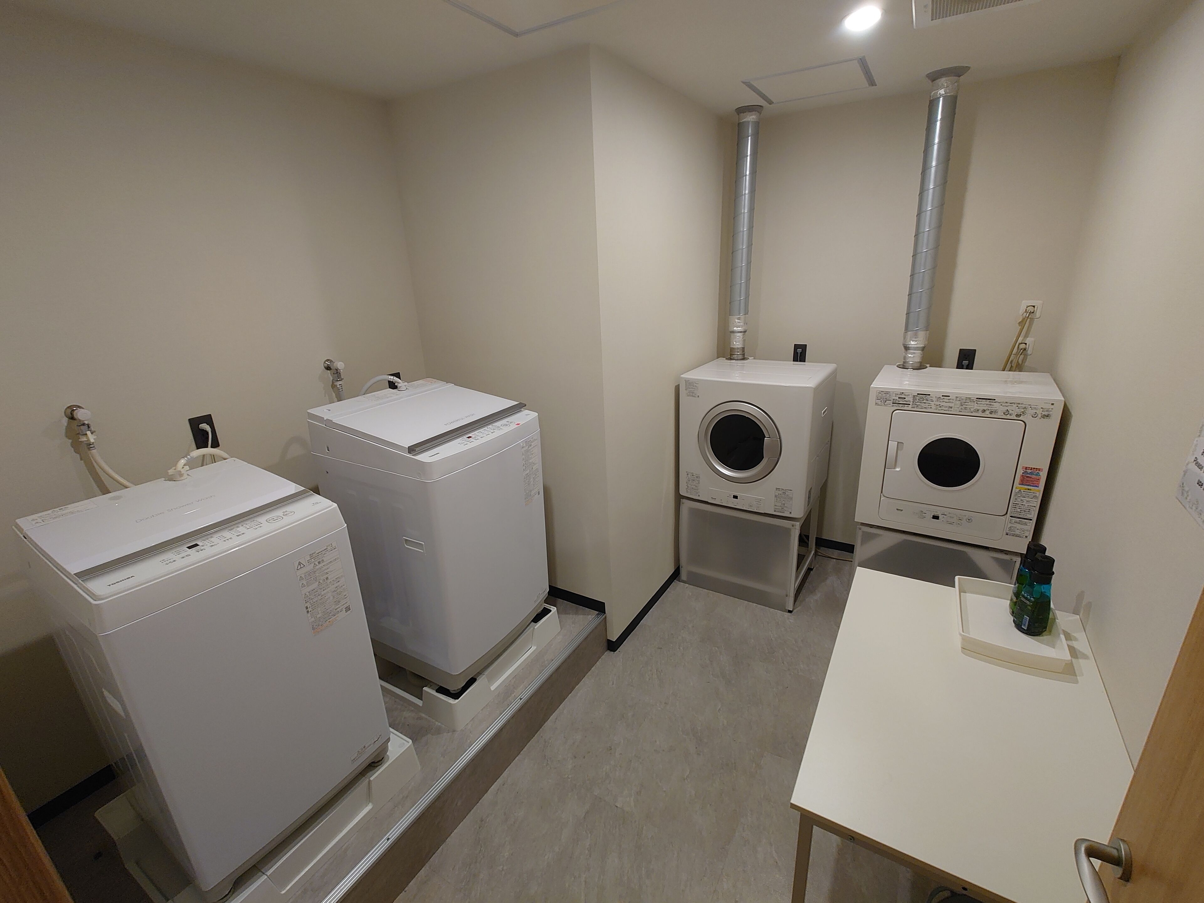 A laundry room with two dryers, a washing machine, and a small table with a bottle.