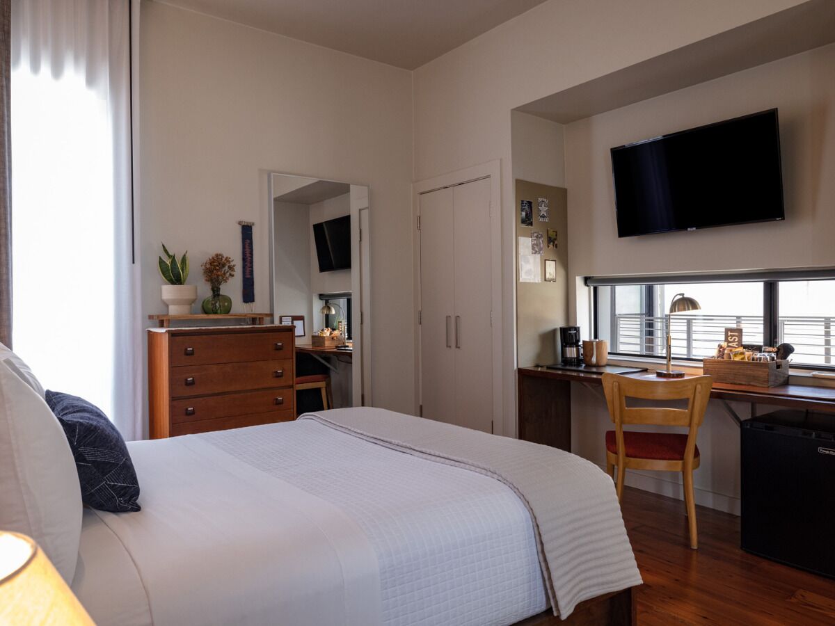 A bedroom with a bed, a desk, a chair, a TV, and a window.