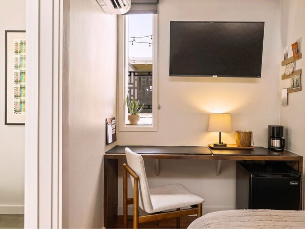 A compact hotel room with a desk, chair, bed, TV, and a small window with a plant.