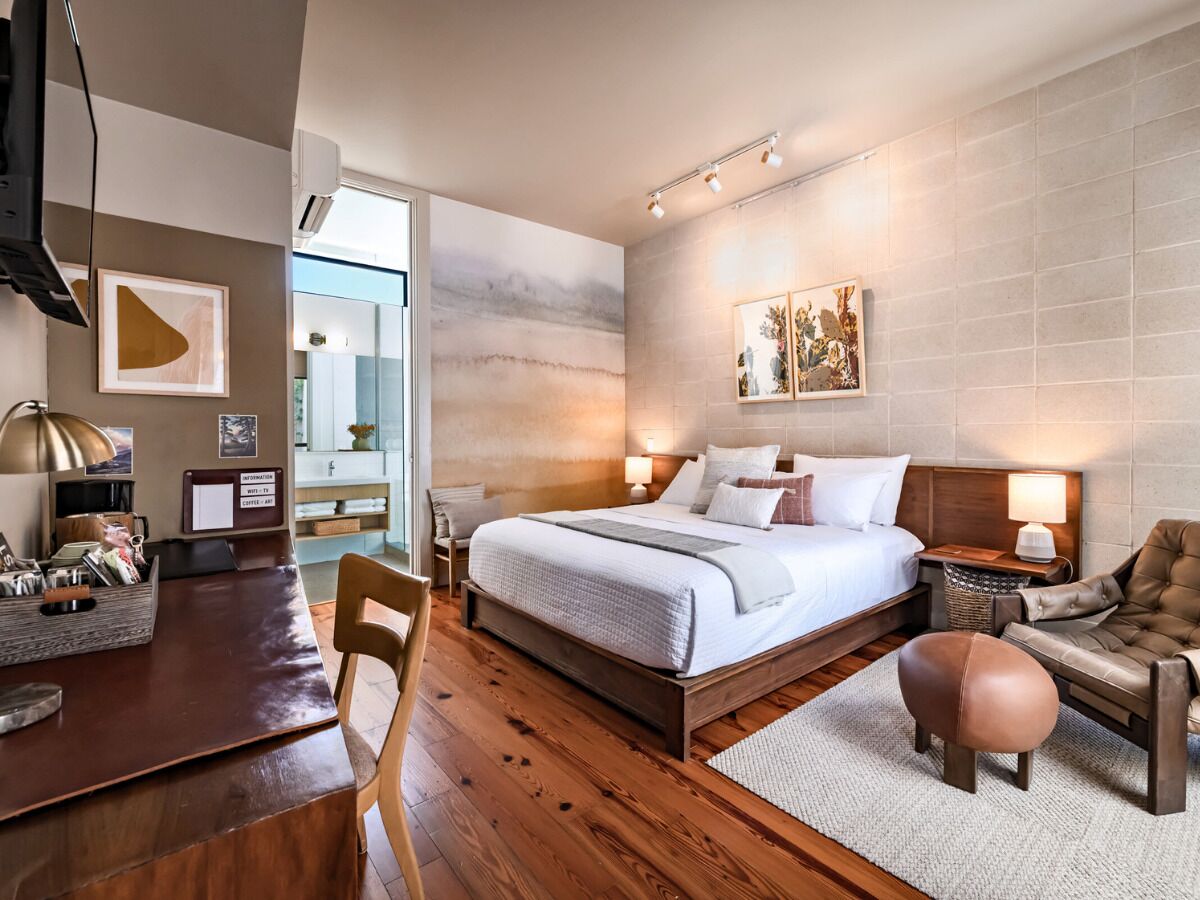 A modern hotel room with a wooden floor, a large bed with white linens, a desk with a lamp, and a chair.
