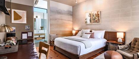 A modern hotel room with a wooden floor, a large bed with white linens, a desk with a lamp, and a chair.
