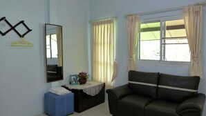 A room with a dark green sofa, a small table with a vase of flowers, a mirror, and a window with curtains. - Bann Suan Rachawadee B&B (Sisaket)
