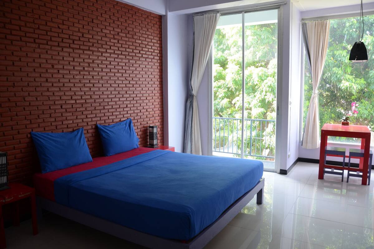 Un dormitorio con pared de ladrillo, una cama grande con ropa de cama azul, una mesita con una silla y vista a la vegetación por la ventana.