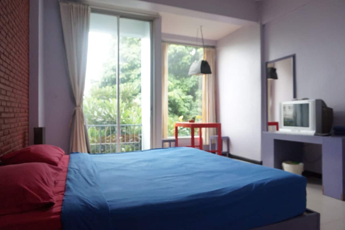 Un dormitorio con cama, televisor, escritorio y ventana con vista a follaje.