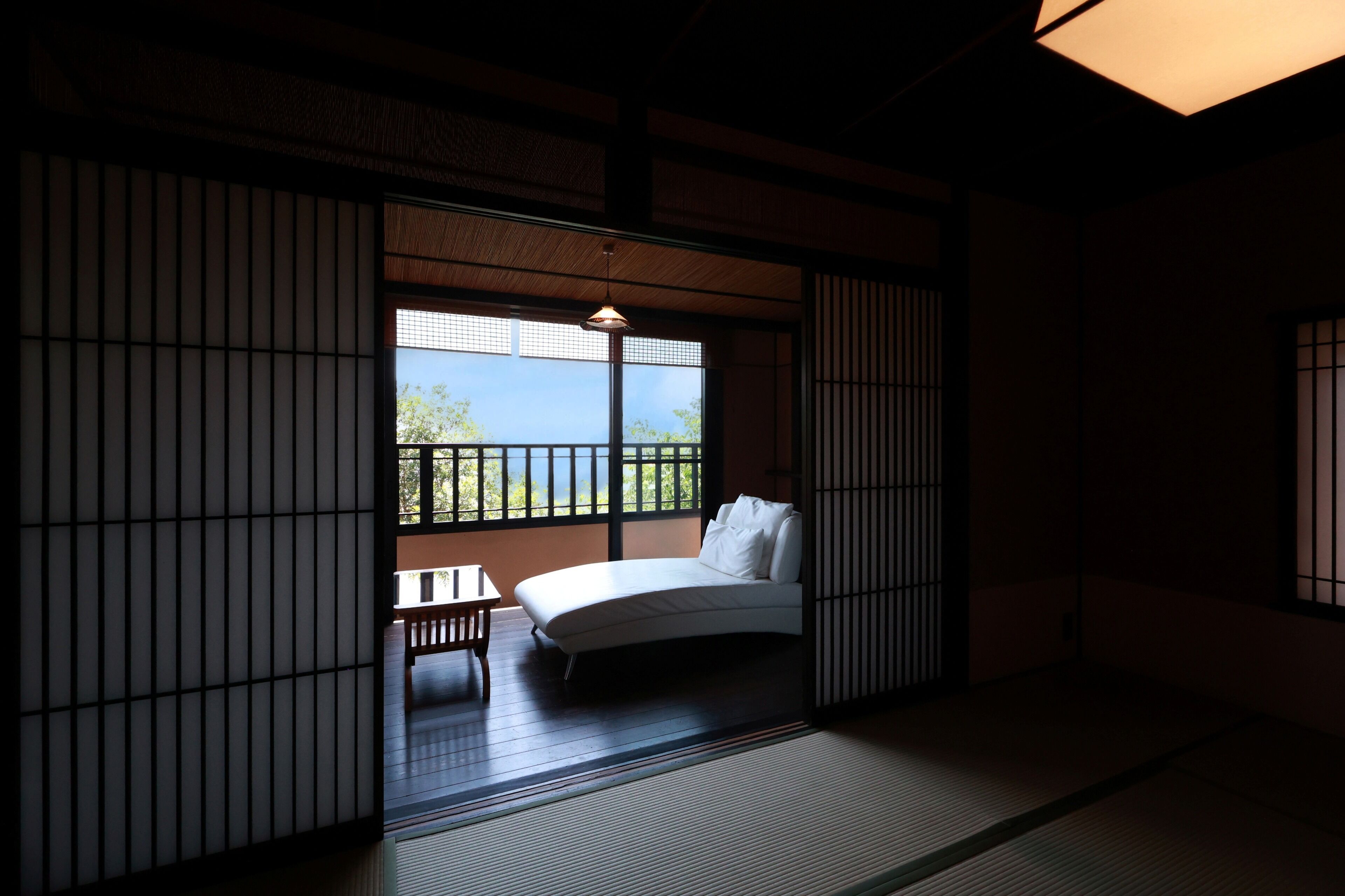 Ett traditionellt japanskt rum med tatamimattor på golvet, en låg säng, ett litet bord och utsikt över grönskan utanför.