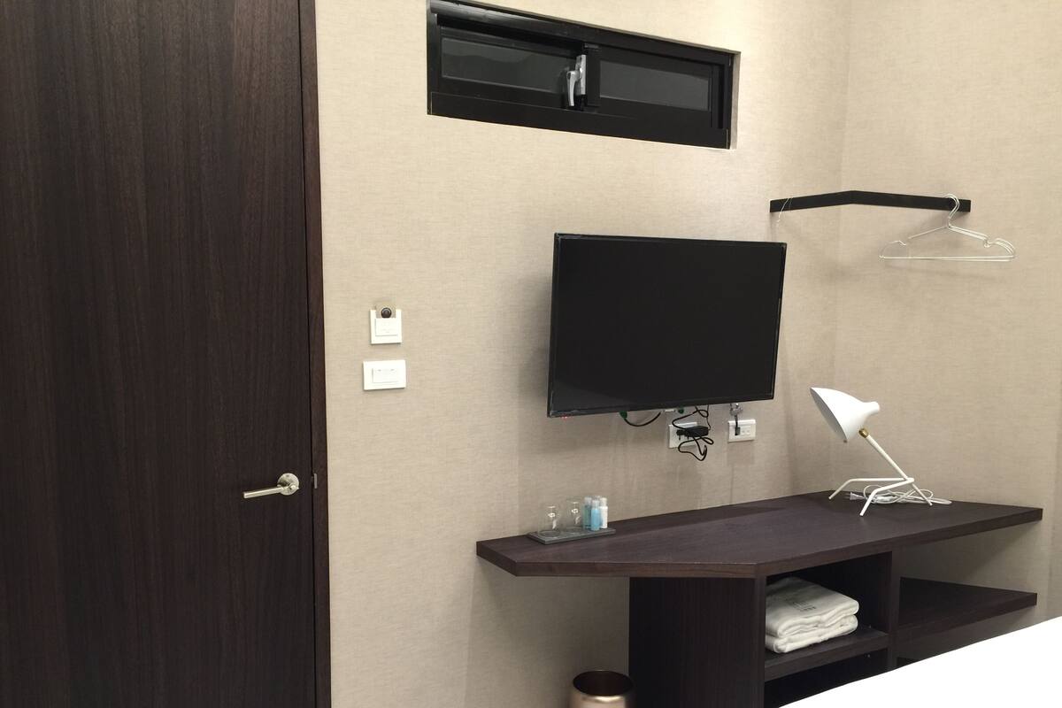 A modern hotel room with a dark wood door, a flat-screen TV mounted on the wall, a white desk lamp, a small shelf with toiletries, and a hanging clothes rack.