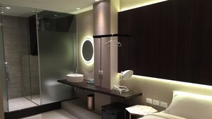 A modern bathroom with a glass shower, a round mirror, a sink, and a bedside table with a lamp. - Feliz Hotel Kenting (Hengchun)