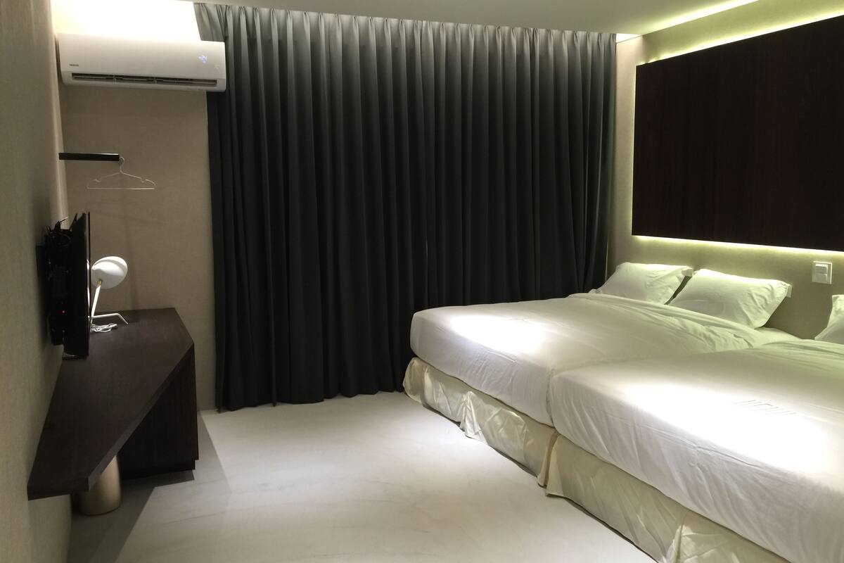 A hotel room with a bed, a bedside table, a TV, and an air conditioning unit.