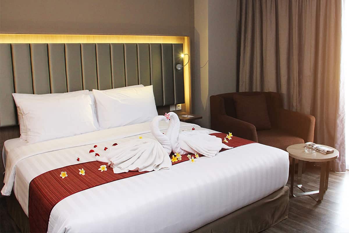 Una habitación de hotel con una cama grande, un sillón marrón, una mesita redonda pequeña y una mesita de noche con lámpara.