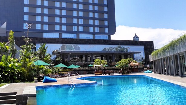 A hotel pool area with a large building in the background, surrounded by greenery and umbrellas.