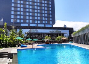 A hotel pool area with a large building in the background, surrounded by greenery and umbrellas. - Gammara Hotel Makassar (Makassar)