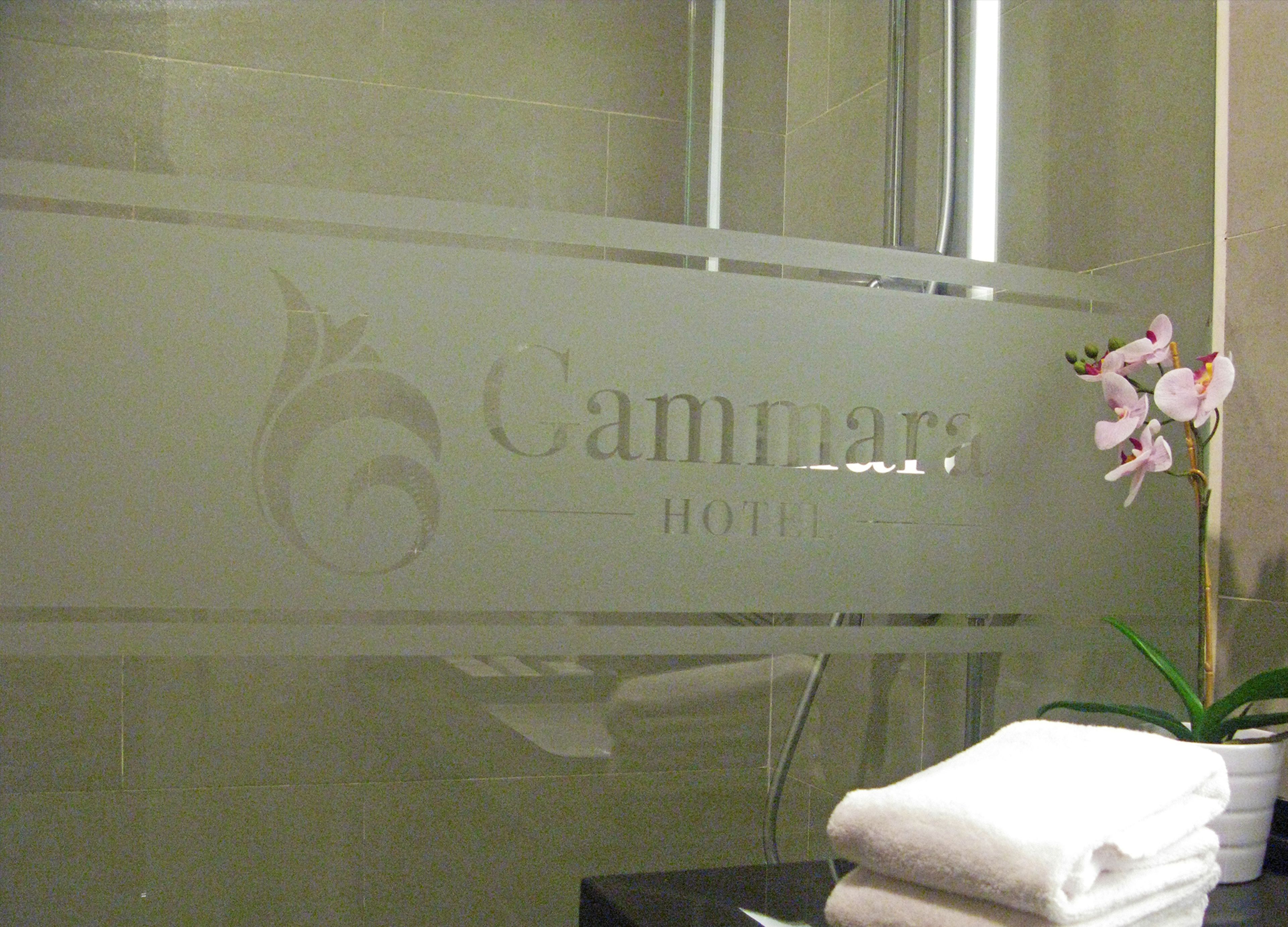 A bathroom with a glass partition displaying the name 'Cammars Hotel'. There is a potted orchid and a stack of white towels visible.