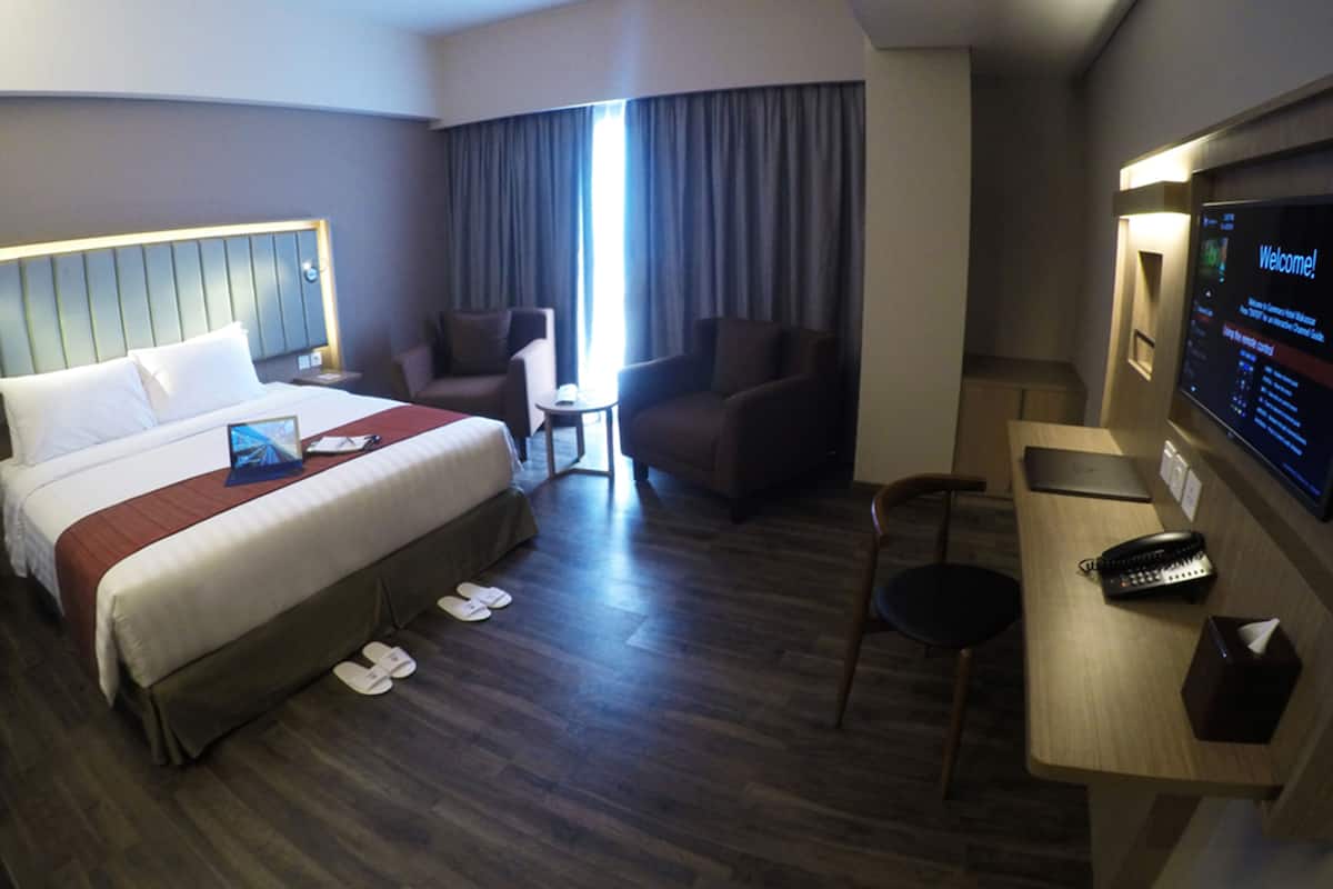 Habitación de hotel con cama, dos sillones, un escritorio con computadora y televisor mostrando '¡Bienvenido!'.