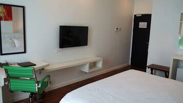 Une chambre d’hôtel avec un lit, un téléviseur fixé au mur, une petite étagère blanche, une chaise verte et un banc en bois.