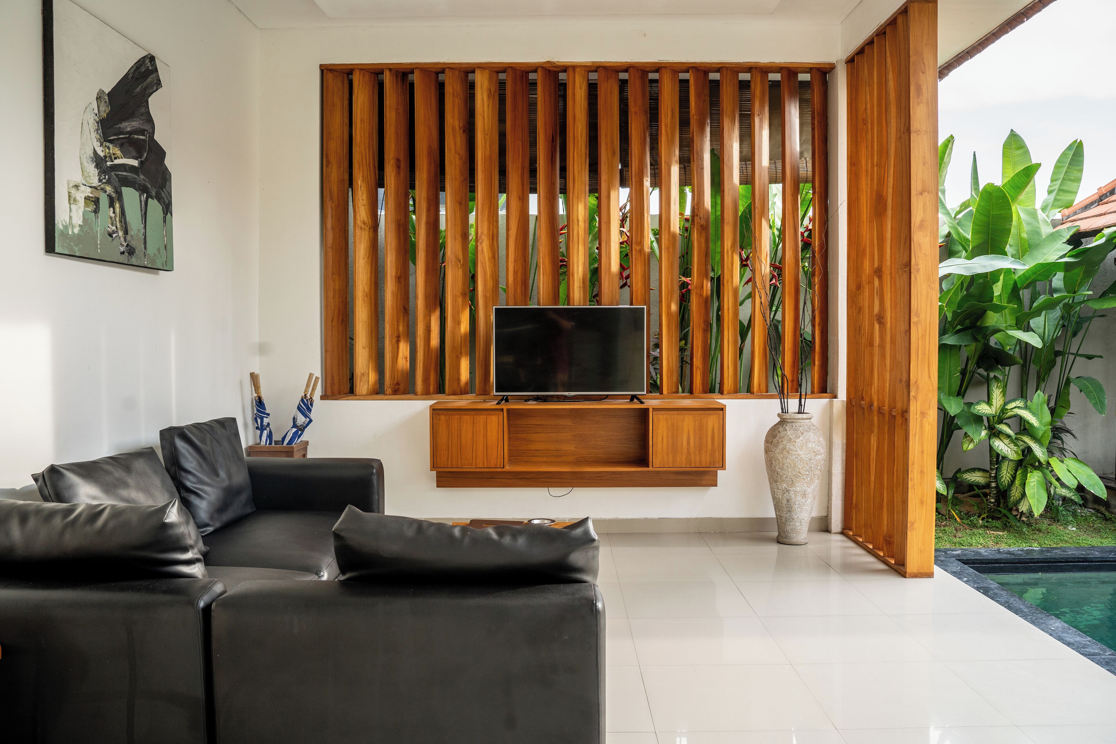 Un salon moderne avec un canapé en cuir noir, un meuble TV en bois et une grande fenêtre équipée de stores verticaux.