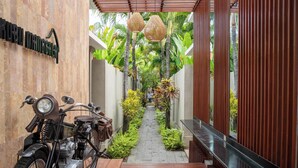 Un étroit sentier bordé d’une végétation luxuriante et d’une moto ancienne mène à un bâtiment moderne doté d’une façade en bois et d’une enseigne indiquant « KUBU MANGCHEP ».
