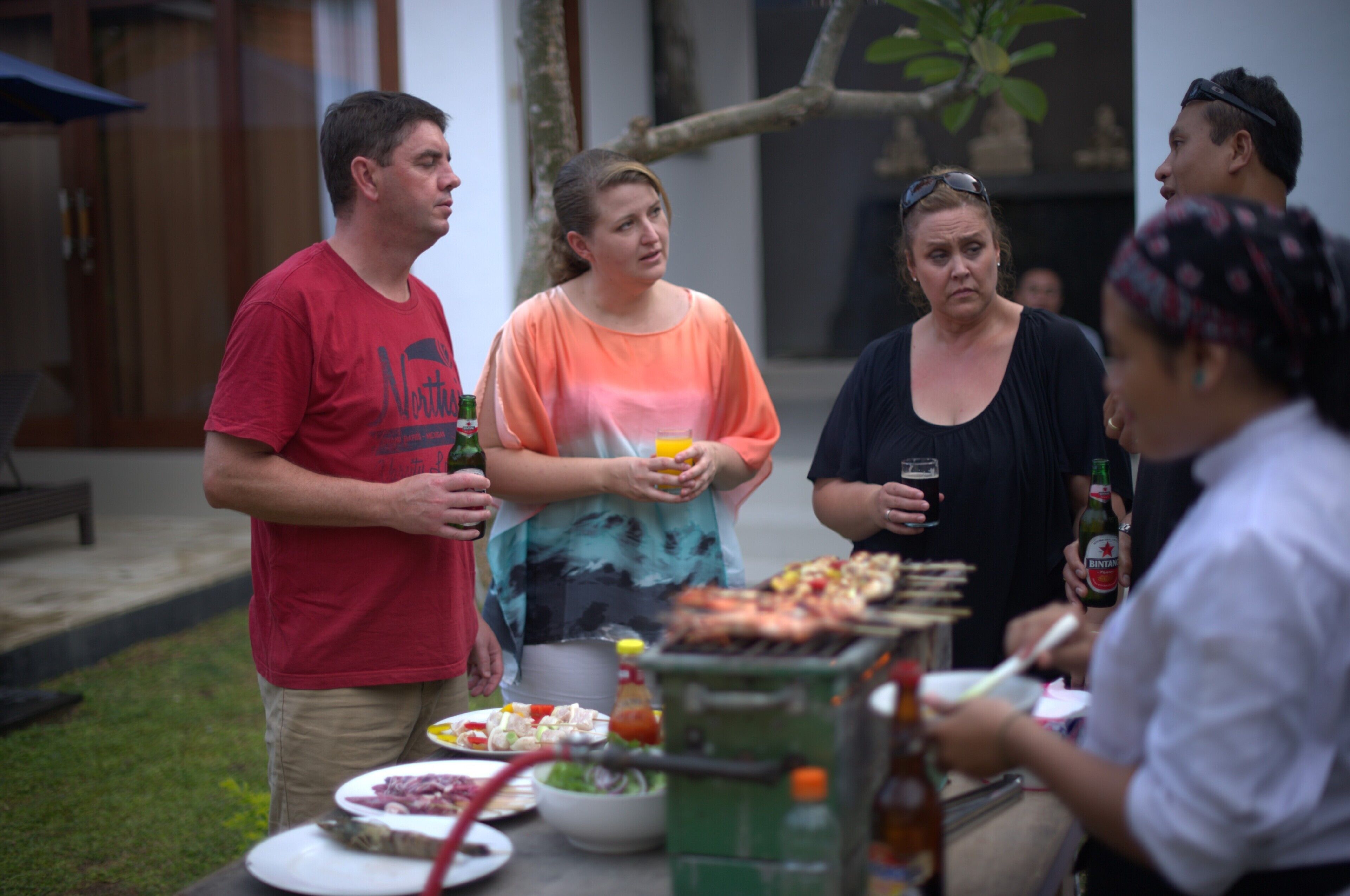 A group of people gathered around a barbecue grill outdoors, with food and drinks on a table.