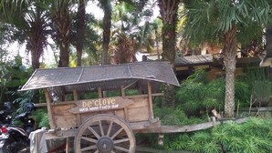 Un carrito de madera rústico con un cartel que dice "De'clove Guest House & Coffee Shop" estacionado en un área pavimentada, con exuberante vegetación y palmeras al fondo.