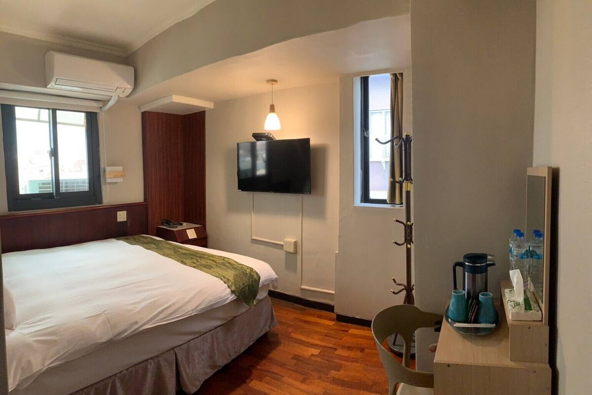 A hotel room with a bed, a TV mounted on the wall, a window with curtains, a wooden floor, and a desk with a kettle and cups.
