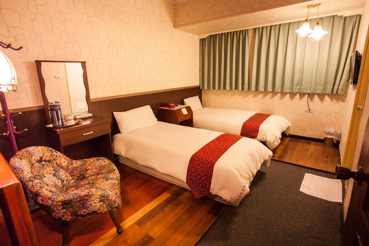 A hotel room with two beds, a chair, a desk, and a window with curtains.