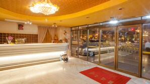 Zona de recepción de un hotel con piso de mármol, alfombra roja, candelabro y grandes ventanales con vista a la calle.