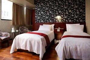 A hotel room with two beds, a floral wallpaper, a nightstand, a chair, and a window with curtains.