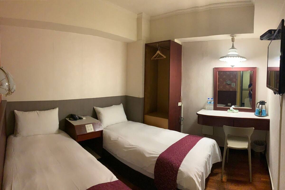 A hotel room with two single beds, a desk with a chair, a mirror, and a hanging coat rack.