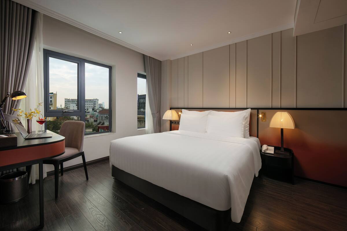 Une chambre d’hôtel avec un grand lit, deux lampes de chevet, un bureau avec un ordinateur portable et une vue sur la ville par une grande fenêtre.