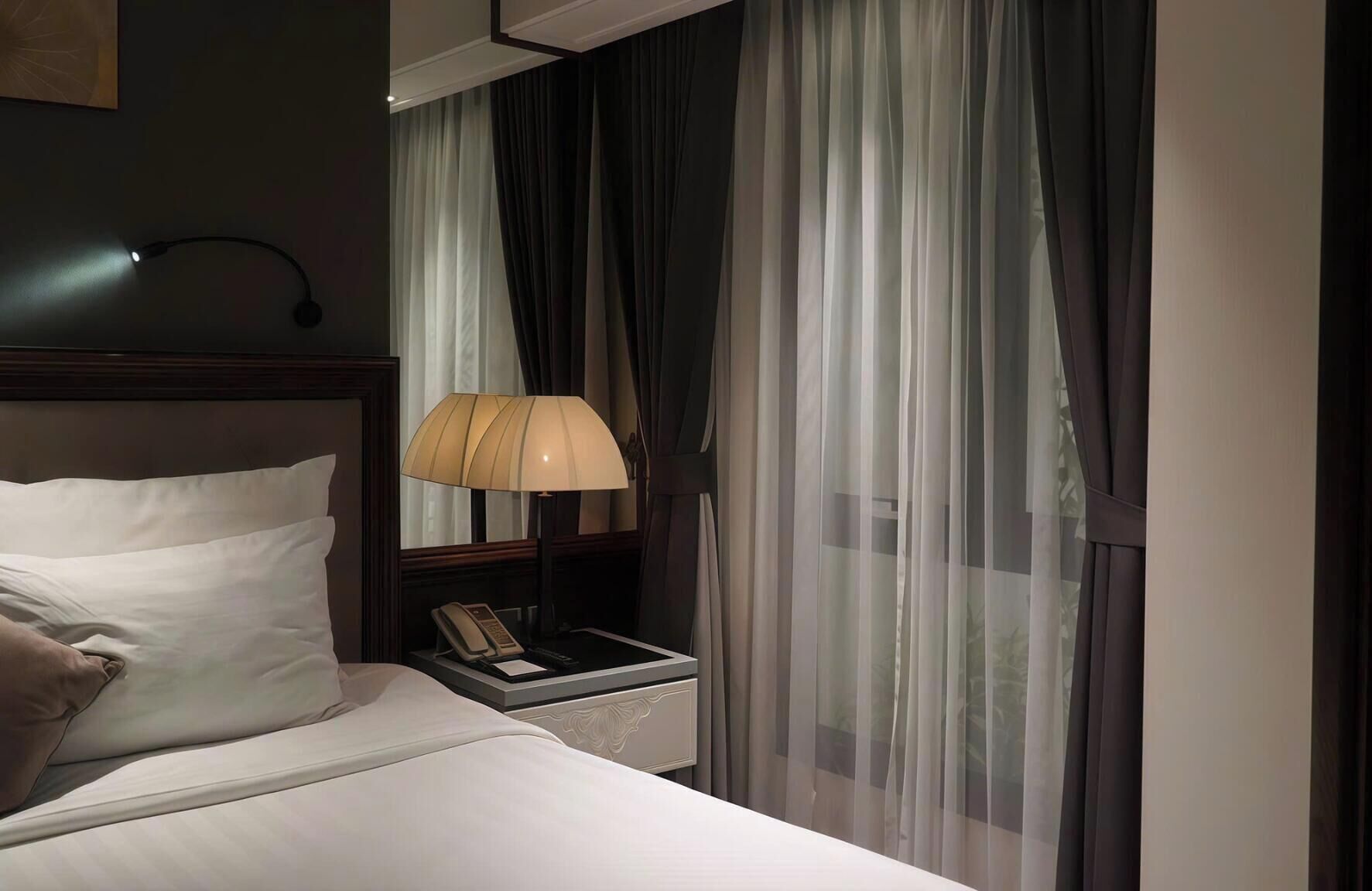 A hotel room with a bed, a bedside lamp, a nightstand with a phone, and sheer curtains.