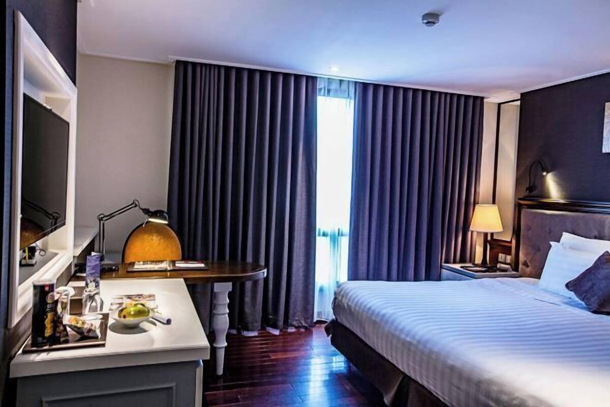 Une chambre d’hôtel avec un lit, un bureau avec une lampe, une chaise, un miroir et une fenêtre avec des rideaux.
