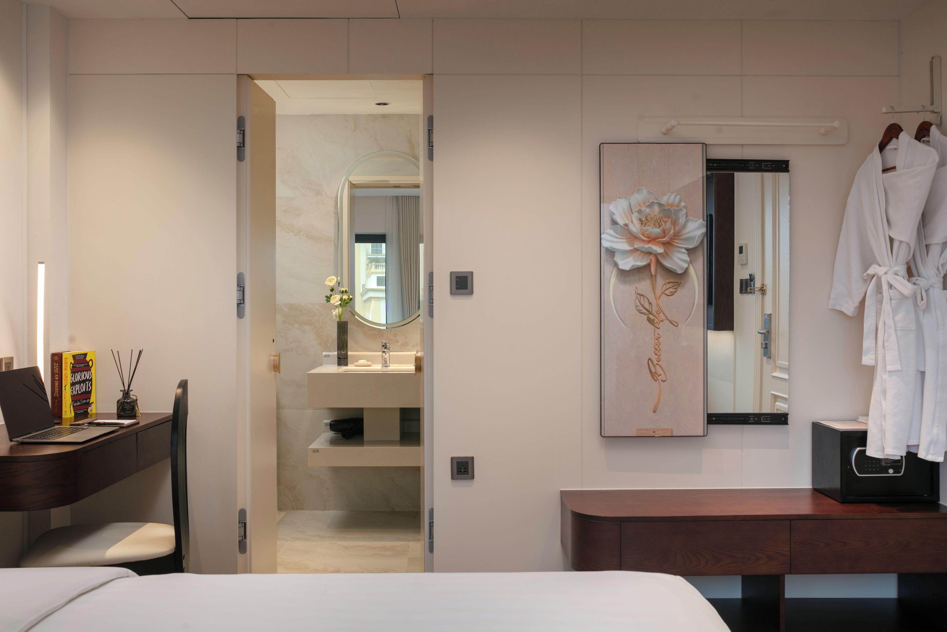 A modern hotel room with a bed, a desk with a laptop, a vanity with a mirror, and a robe rack.