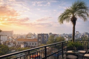 Un balcón con sillas y una palmera, con vista a un paisaje urbano que incluye edificios y una iglesia.