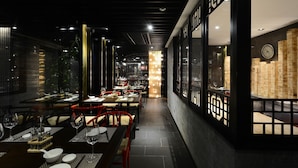 L’intérieur d’un restaurant à la lumière tamisée, avec des tables dressées pour le dîner, un mur décoratif orné d’une horloge et une source de lumière chaleureuse en arrière-plan.