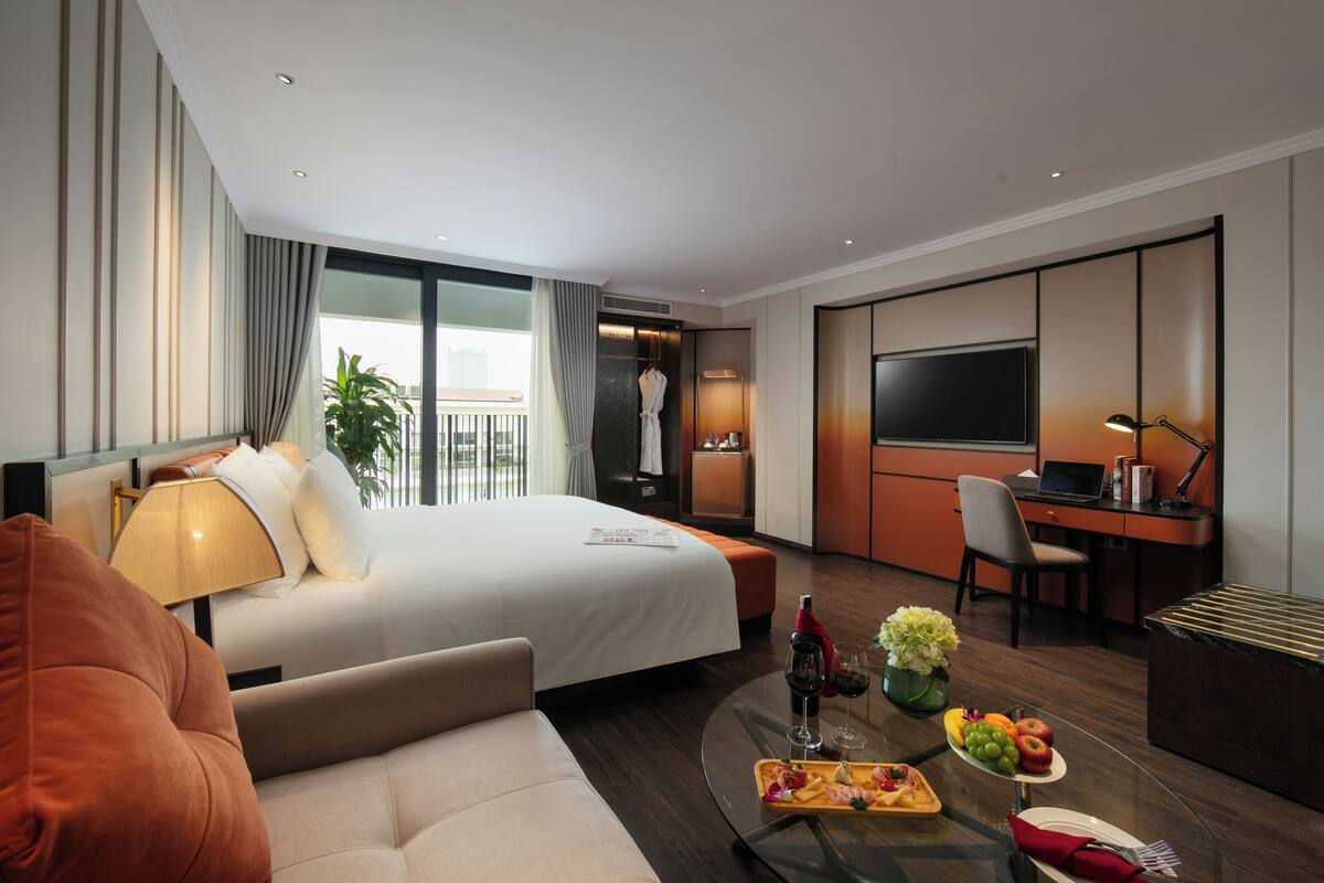 Une chambre d’hôtel moderne dotée d’un grand lit, d’un bureau avec un ordinateur portable, d’une télévision et d’un coin repas avec des fruits et du vin.