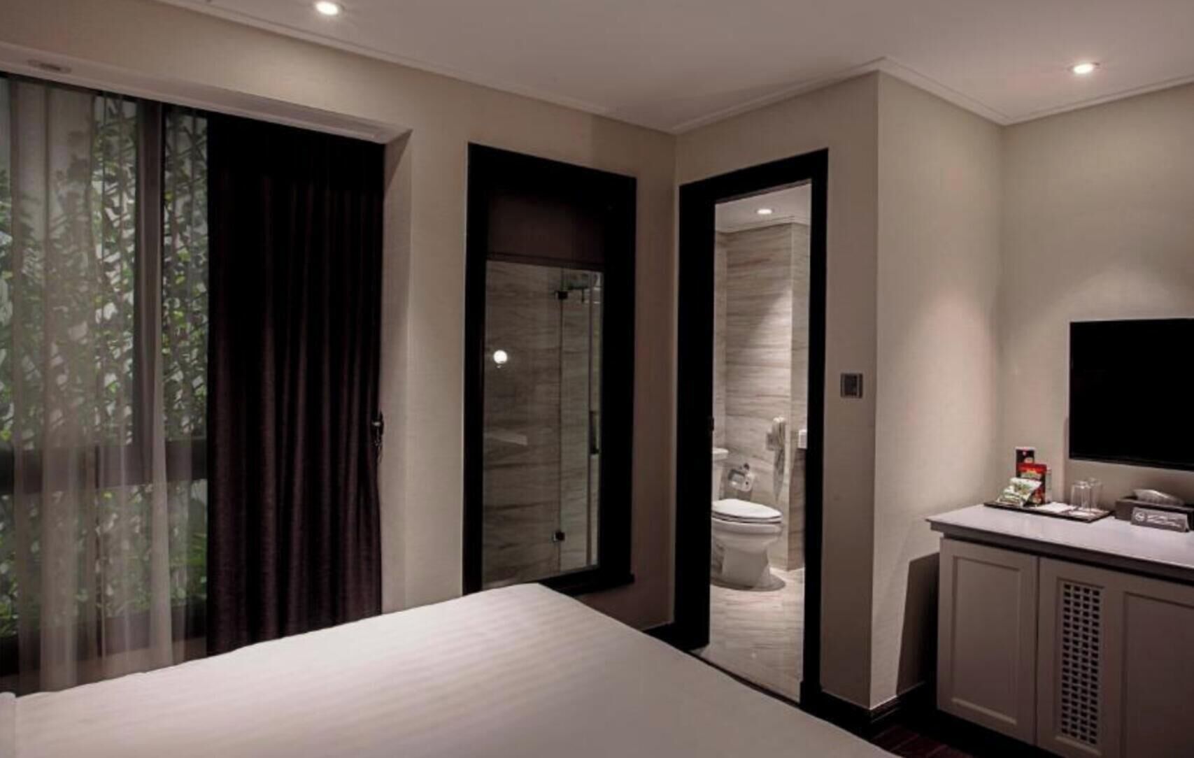 Habitación de hotel con cama, escritorio, televisor, baño con inodoro y puerta corrediza de vidrio a un balcón.
