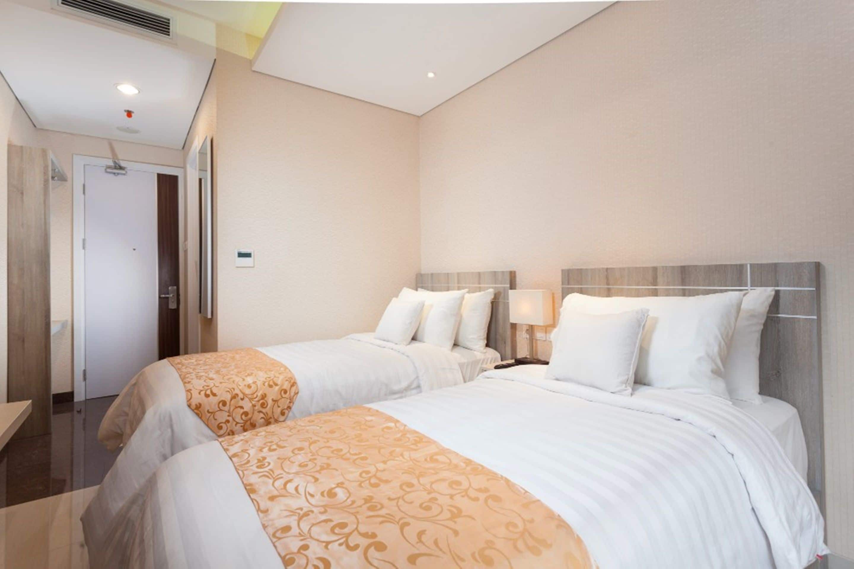 A hotel room with two beds, a wooden headboard, a bedside table with a lamp, and a door leading to a bathroom.