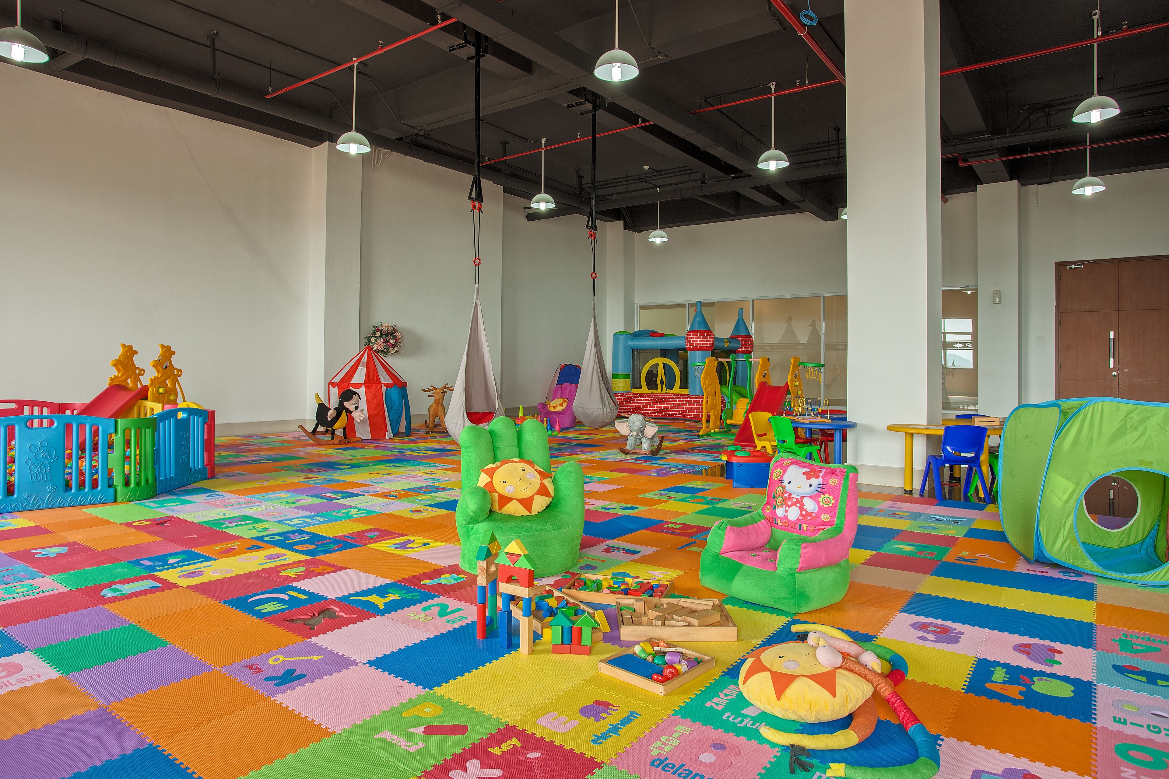 Un espace de jeux intérieur, bien éclairé, avec des tapis de jeu colorés, une structure de jeu verte et divers jouets.