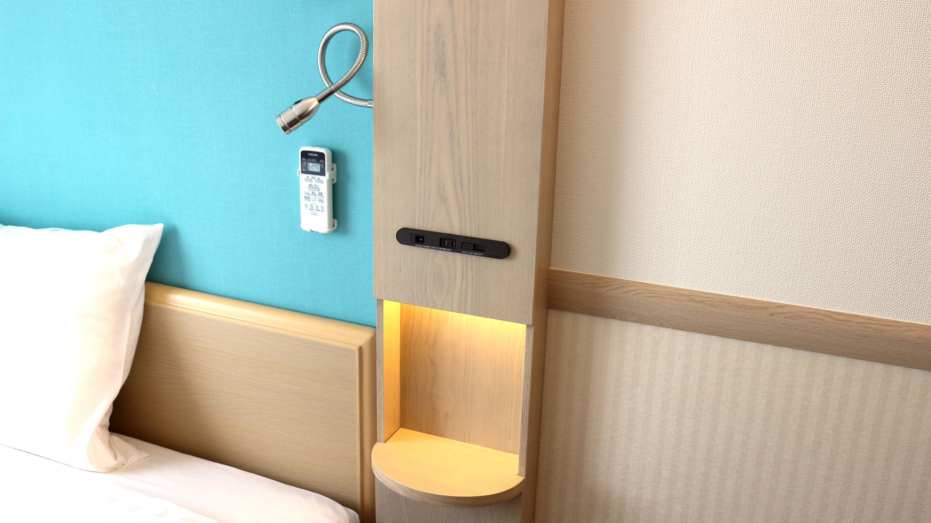 A hotel room with a bed, a bedside table, a remote control, and a wall-mounted device.