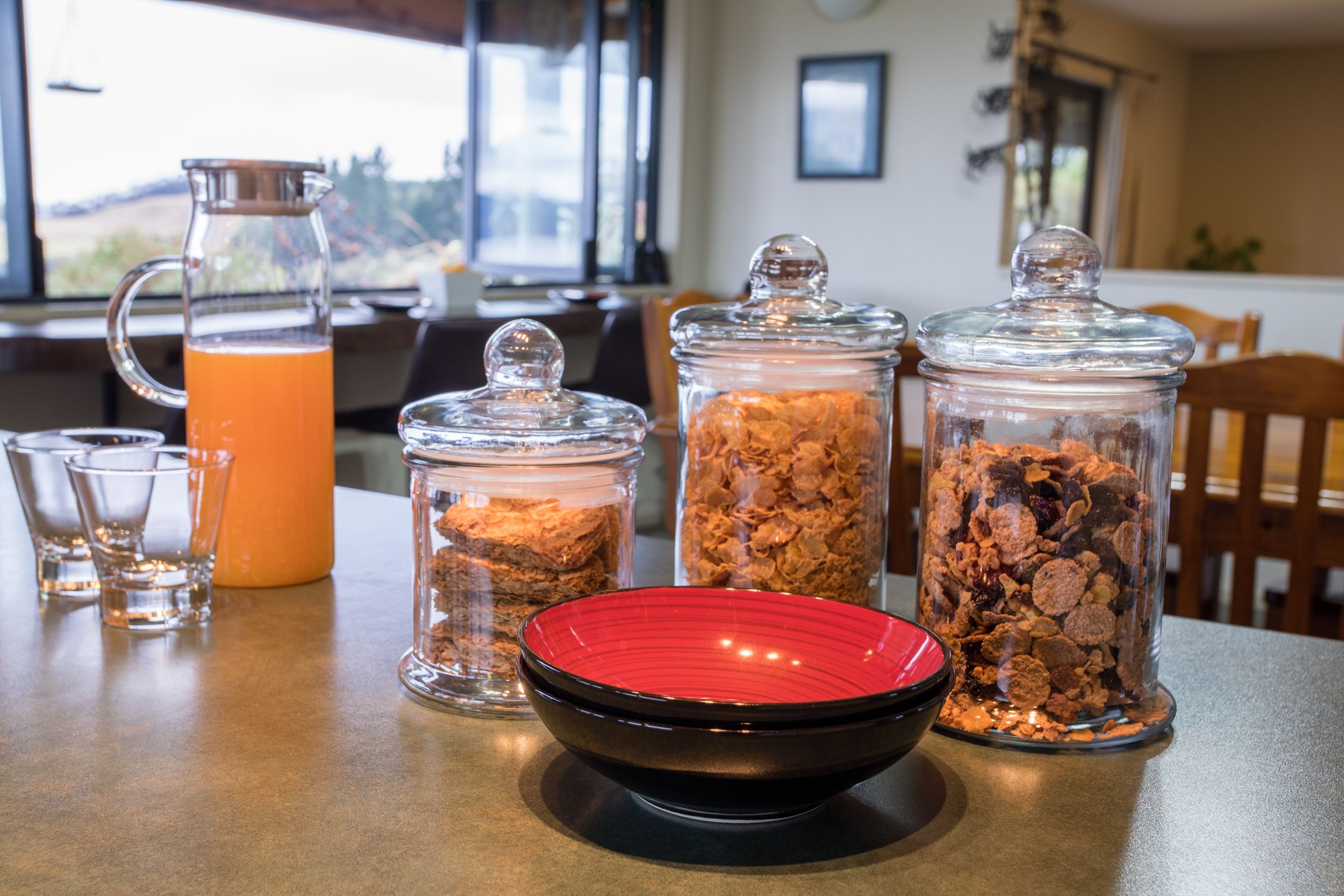 A breakfast table with a pitcher of orange juice, two glass jars with granola, and a red bowl.