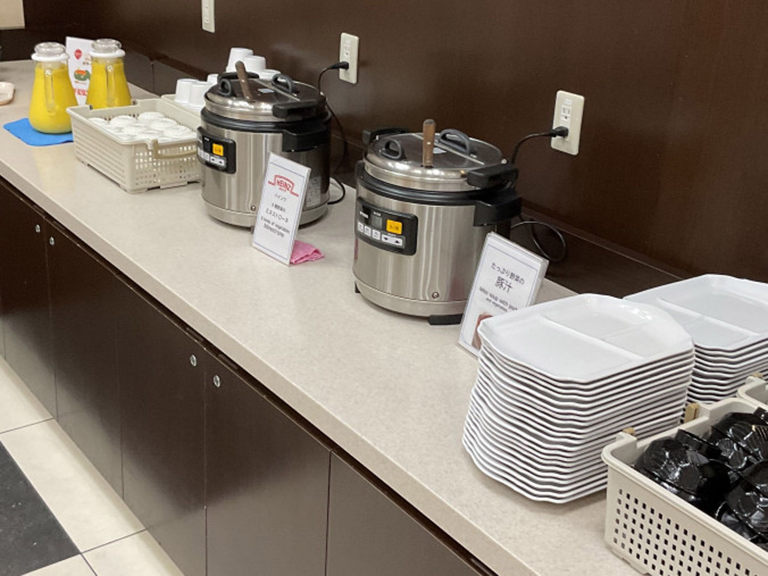 A breakfast buffet setup with hot food containers, juice dispensers, and stacked plates.