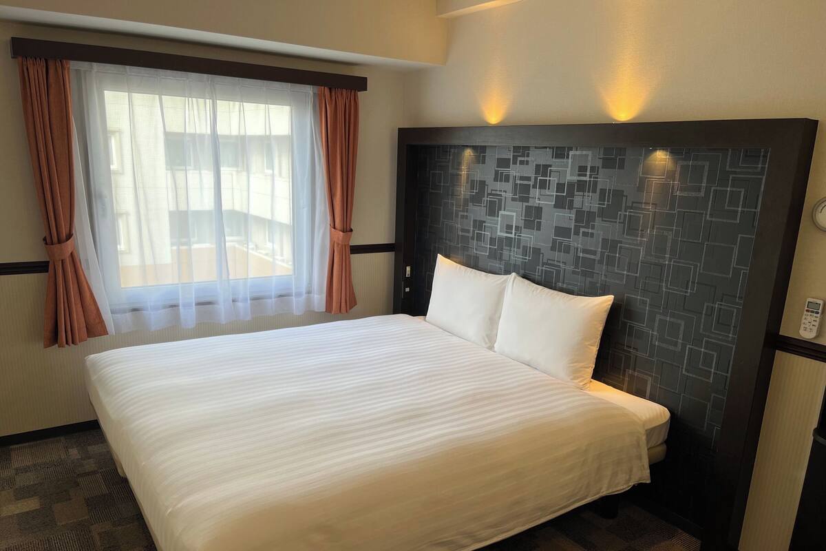 A hotel room with a large bed, a headboard with a geometric pattern, a window with sheer curtains, and a wall-mounted phone.