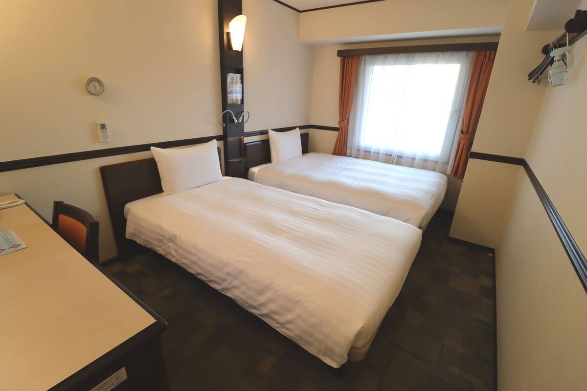 A hotel room with two beds, a desk, a chair, a clock, and a window with curtains.