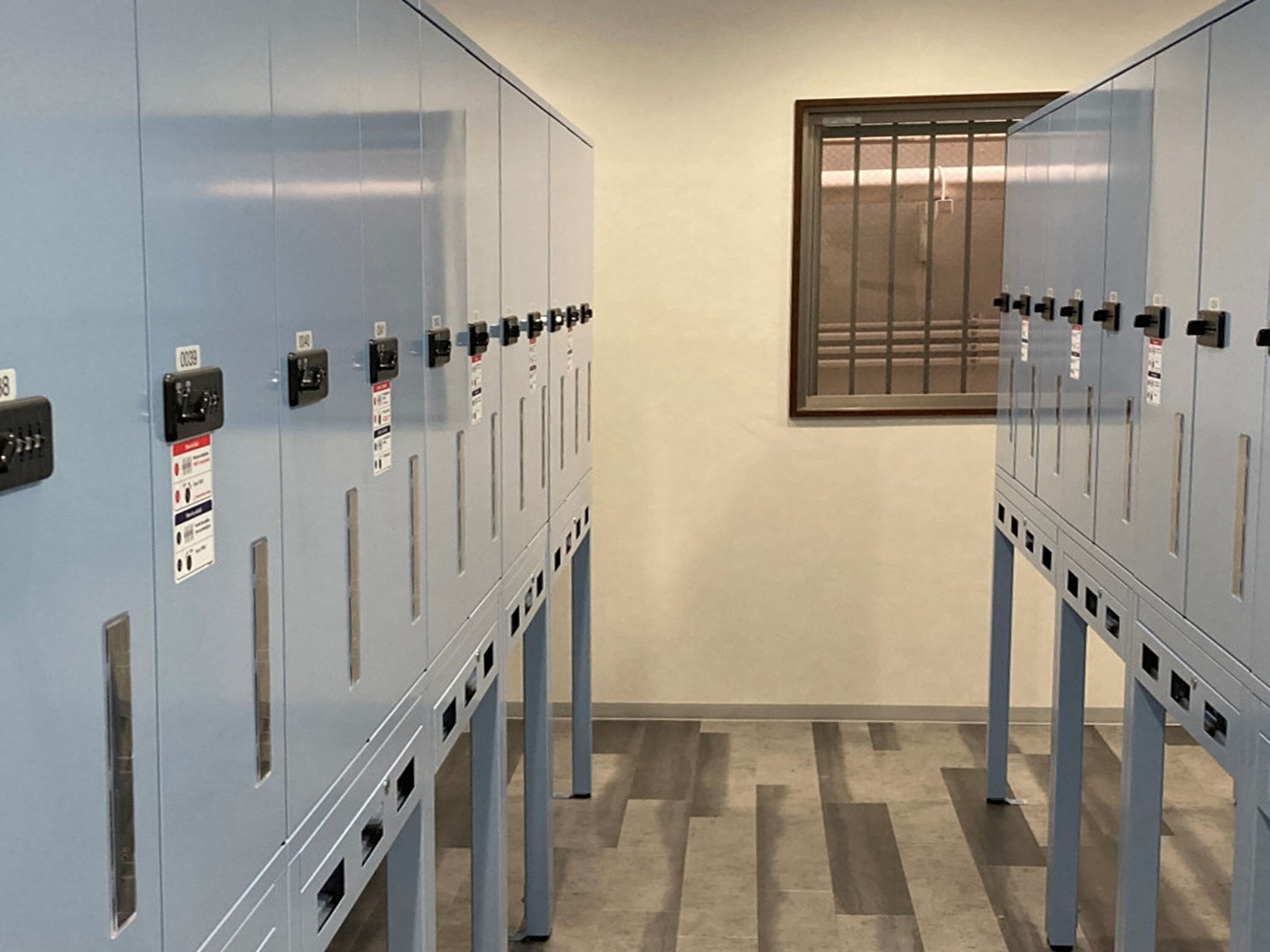 A row of blue lockers with numbered labels, a window with bars, and a tiled floor.