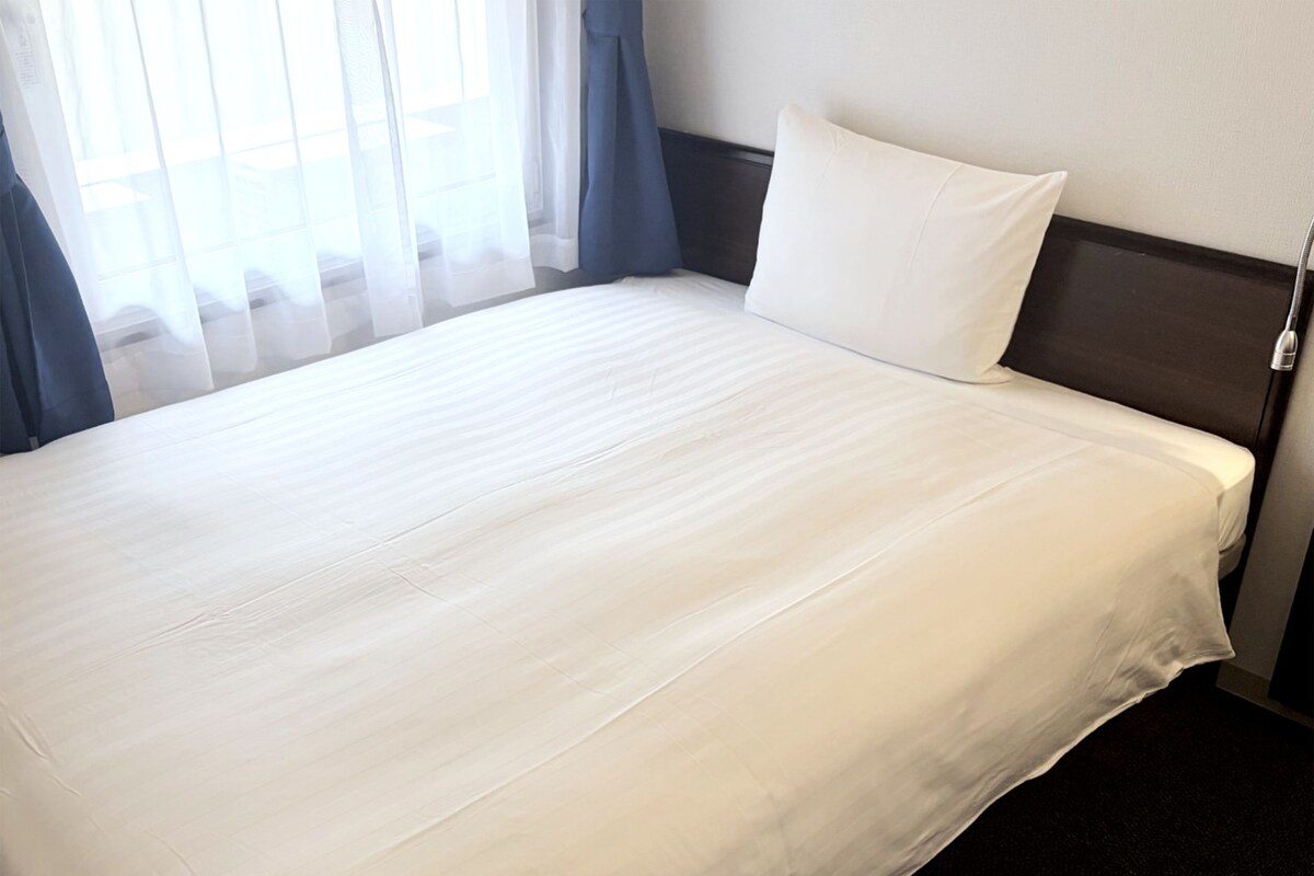 Un lit bien fait, avec un drap blanc et un oreiller, une tête de lit sombre et une fenêtre ornée de rideaux légers.