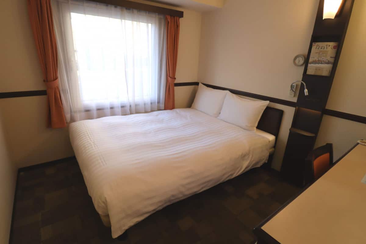 A hotel room with a bed, a window with curtains, a bedside table, and a wall clock.