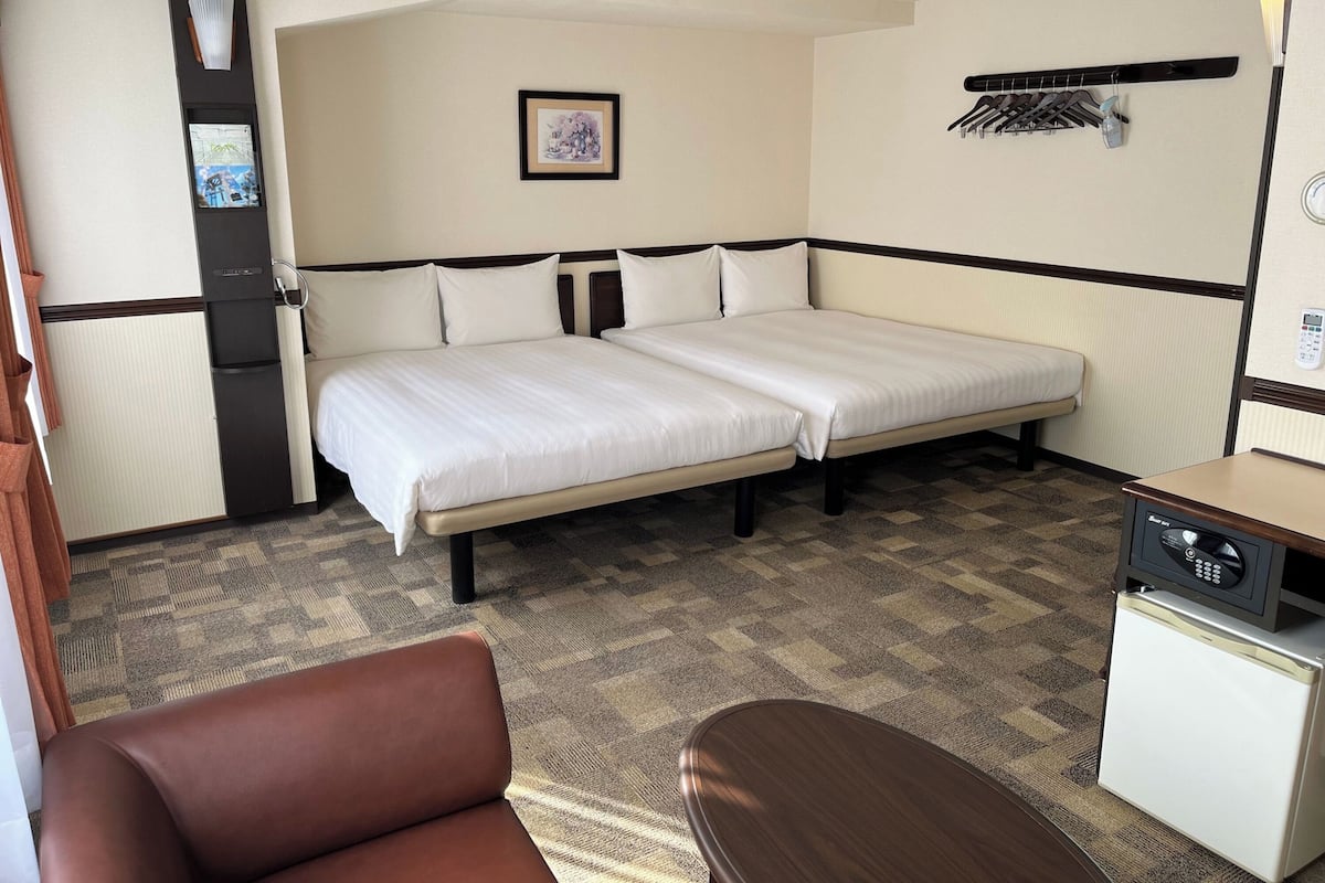 A hotel room with a double bed, a small desk with a refrigerator, a chair, and a wall-mounted coat rack.