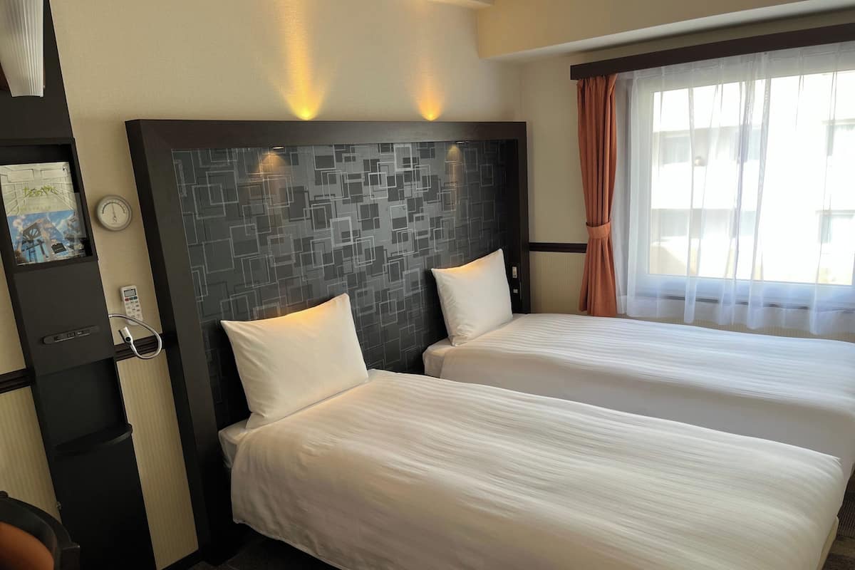 A hotel room with two beds, a modern headboard, a desk with a telephone, and a window with curtains.